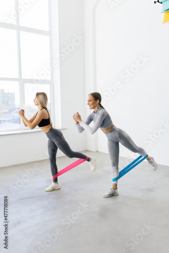 Two smiling athletic women squatting synchronously with outfit in fitness studio. Women are enjoying leg and buttocks work out with resistance band. Training together with friend in gym concept
