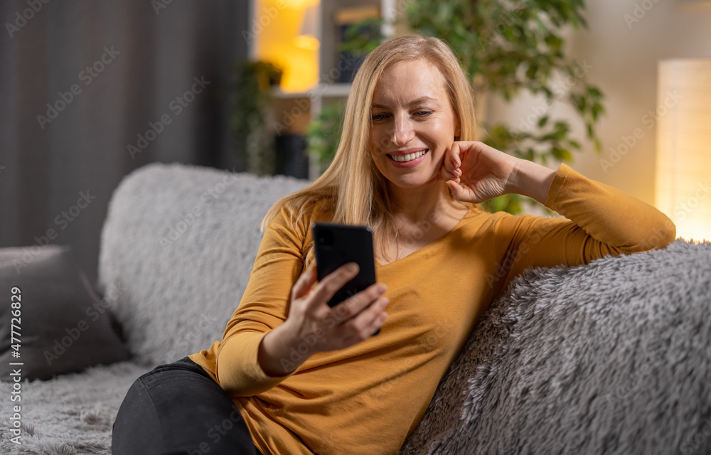 Happy mature woman in casual wear sitting on comfy grey couch and using smartphone. Pretty caucasian lady with blond hair browsing internet on modern gadget. Happy mature woman in casual wear sitting on comfy grey couch and using smartphone. Pretty caucasian lady with blond hair browsing internet on modern gadget.