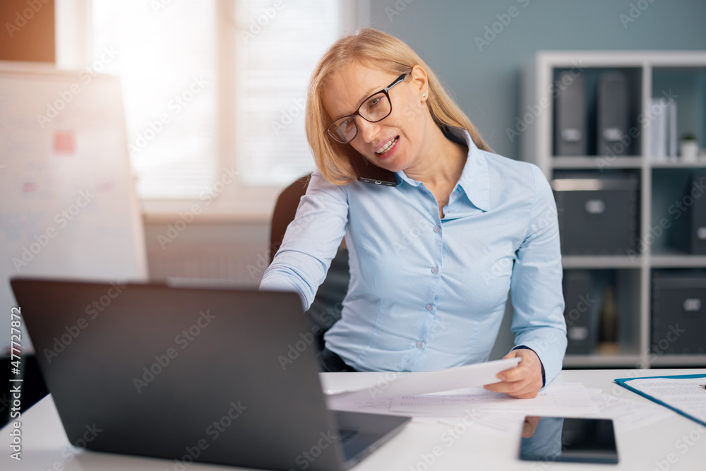 Obraz premium Confident mature woman in eyeglases and blue shirt typing on wireless laptop and talking on mobile phone. Busy lady with blond hair having lots of work to do at office.