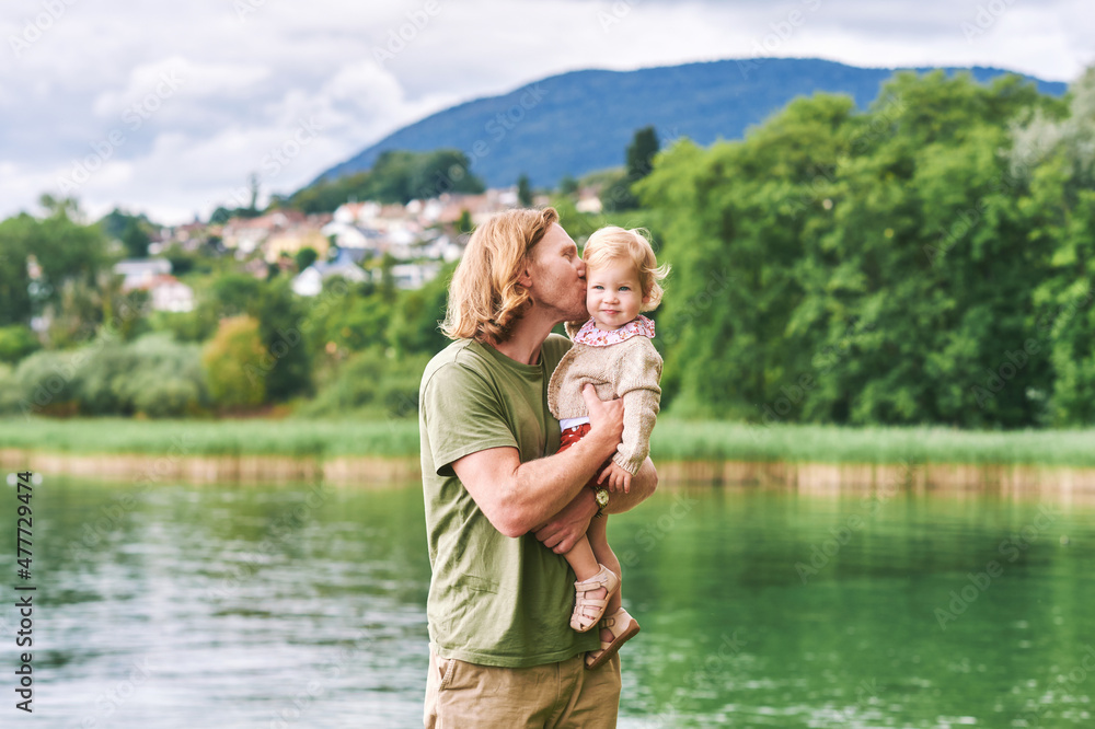 Fototapeta premium Portrait of happy young father with adorable toddler girl playing outside next to lake or river