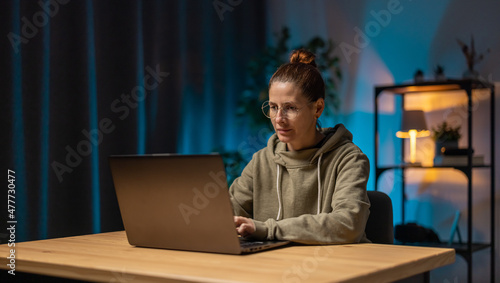 Focused mature woman in eyewear sitting at home office during evening time and working on modern laptop. Freelance, people and technology concept.