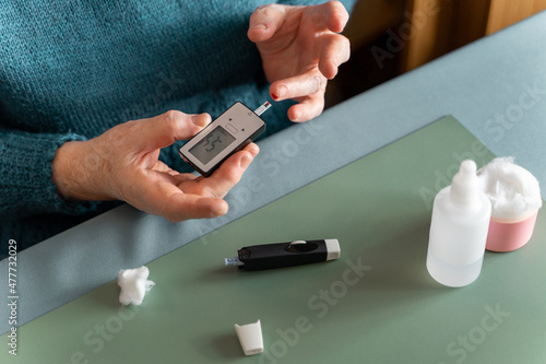 An elderly woman measures her blood sugar level at home with a blood glucose meter. Cotton wool  lancet device and hydrogen peroxide are on the table.