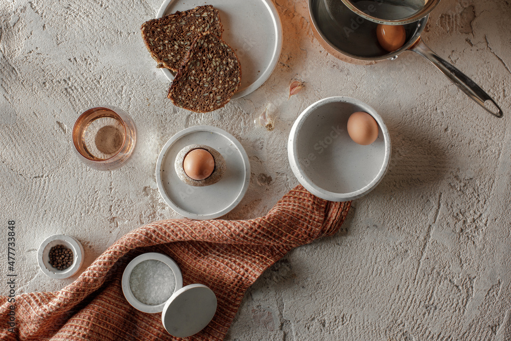 Breakfast setting with boiled egg in stoneware egg cup, whole grain rye bread in concrete tray, salt flakes and pepper in concrete bowls on rough textured clay background. Healthy breakfast concept Breakfast setting with boiled egg in stoneware egg cup, whole grain rye bread in concrete tray, salt flakes and pepper in concrete bowls on rough textured clay background. Healthy breakfast concept