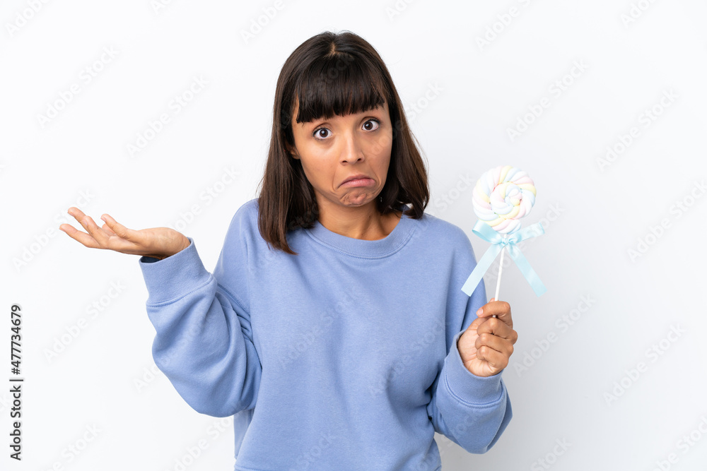 Young mixed race woman holding a lollipop isolated on white background having doubts while raising hands Young mixed race woman holding a lollipop isolated on white background having doubts while raising hands