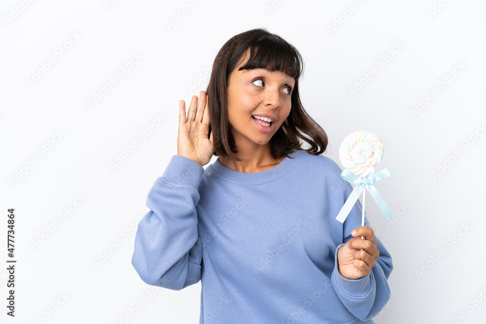 Young mixed race woman holding a lollipop isolated on white background listening to something by putting hand on the ear Young mixed race woman holding a lollipop isolated on white background listening to something by putting hand on the ear