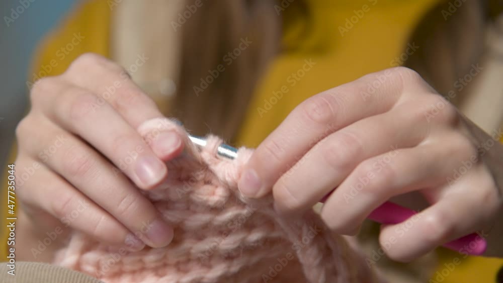 Close-up. Hands of an attractive young caucasian woman in casual clothes sitting on a sofa indoors knits a woolen product by a crochet Close-up. Hands of an attractive young caucasian woman in casual clothes sitting on a sofa indoors knits a woolen product by a crochet