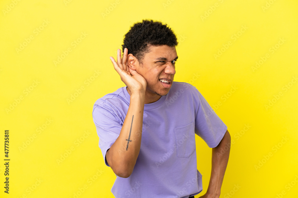 Young African American man isolated on yellow background listening to something by putting hand on the ear Young African American man isolated on yellow background listening to something by putting hand on the ear