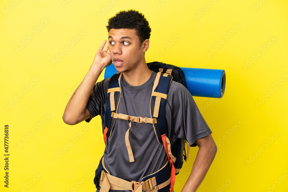 Young mountaineer African American man with a big backpack isolated on yellow background listening to something by putting hand on the ear Young mountaineer African American man with a big backpack isolated on yellow background listening to something by putting hand on the ear