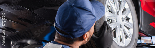African american mechanic in cap looking at car wheel in garage, banner. © LIGHTFIELD STUDIOS