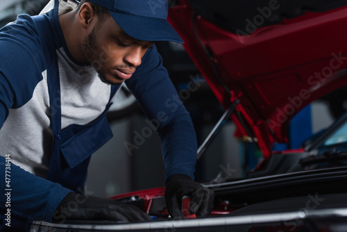 african american technician in uniform and work gloves inspecting car in workshop.