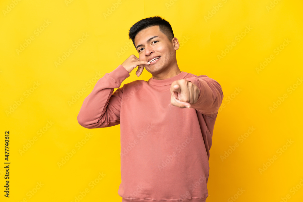 Young Ecuadorian man isolated on yellow background making phone gesture and pointing front Young Ecuadorian man isolated on yellow background making phone gesture and pointing front