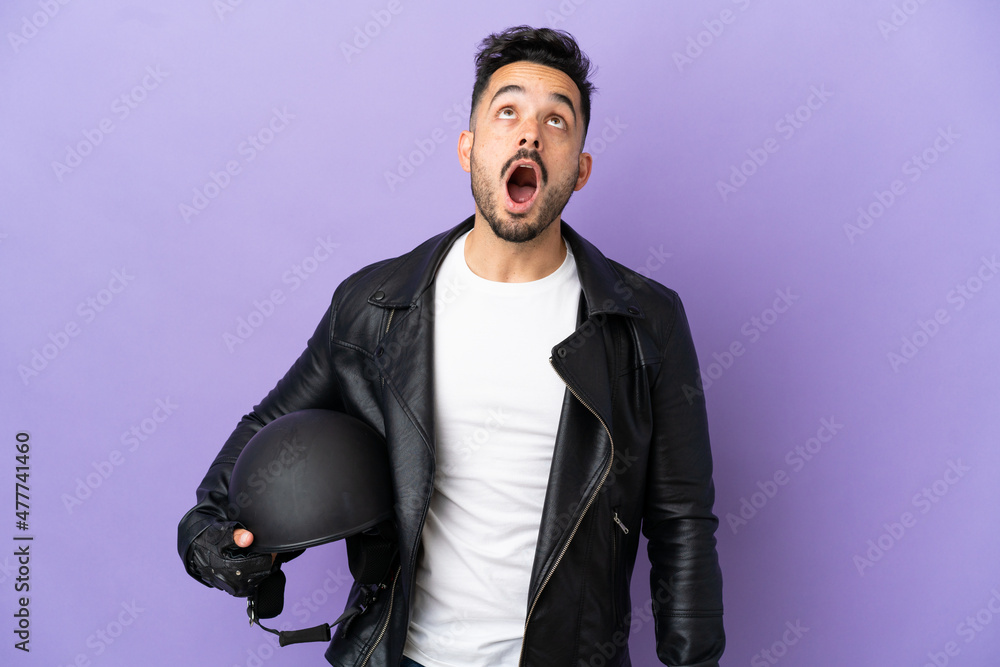 Young man with a motorcycle helmet isolated on purple background looking up and with surprised expression Young man with a motorcycle helmet isolated on purple background looking up and with surprised expression