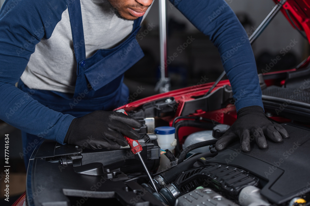 partial view of african american technician in work gloves inspecting car engine compartment with screwdriver.