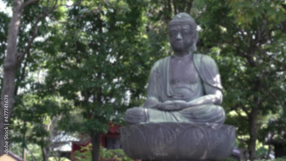Defocused shot of a statue Buddha with green hedges and trees. Peaceful scene in famous ancient buddhist temple Senso-ji. Japanese shinto shrine in Tokyo City, Japan. Blurred background in the scene Defocused shot of a statue Buddha with green hedges and trees. Peaceful scene in famous ancient buddhist temple Senso-ji. Japanese shinto shrine in Tokyo City, Japan. Blurred background in the scene