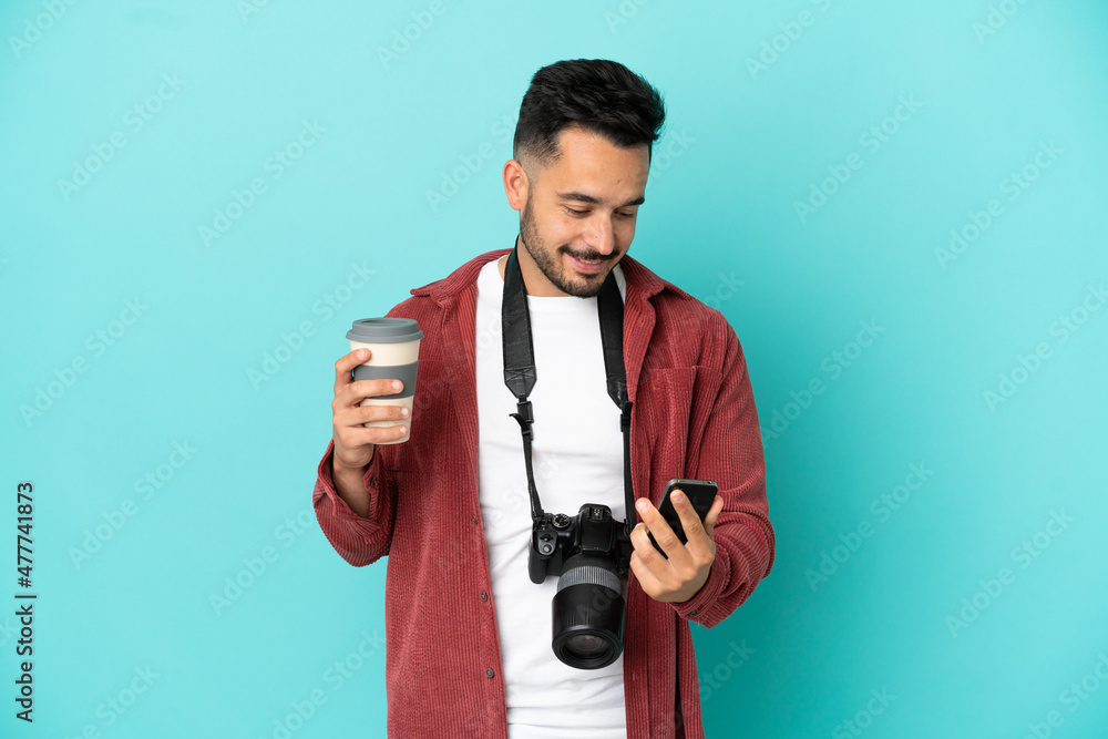 Young photographer caucasian man isolated on blue background holding coffee to take away and a mobile Young photographer caucasian man isolated on blue background holding coffee to take away and a mobile