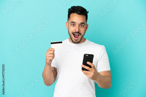 Young caucasian man isolated on blue background buying with the mobile and holding a credit card with surprised expression