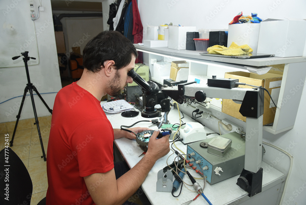 A young man in the workshop examines the microcircuit of an electrical device through a microscope, performing electrical soldering of equipment