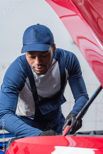 young african american technician in overalls and cap inspecting car with open hood.