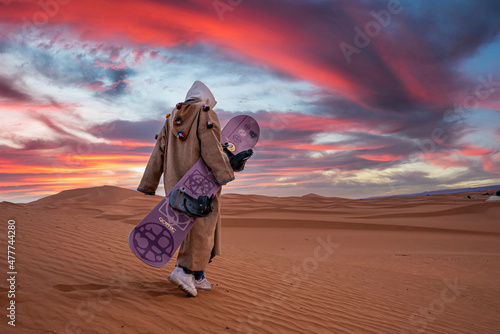 Sahara, Morocco. October 10, 2021. Man in traditional clothes carrying sandboard while walking on sand against cloudy sky during dusk, Man with sandboard walking in desert photo