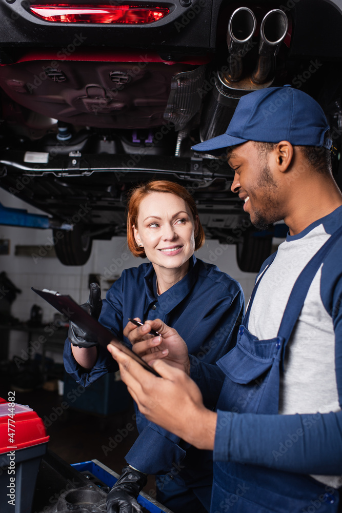 Smiling multiethnic mechanics with clipboard talking near toolbox and car in service. Smiling multiethnic mechanics with clipboard talking near toolbox and car in service.