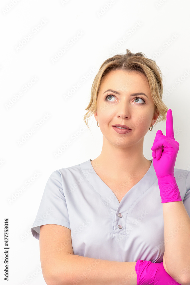 Portrait of young caucasian doctor woman wearing rubber gloves in medical uniform with one finger up, looking up against a white background, copy space Portrait of young caucasian doctor woman wearing rubber gloves in medical uniform with one finger up, looking up against a white background, copy space