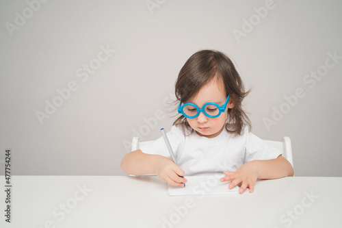 the child plays doctor. a girl in toy blue glasses sits at a white table. space for text and advertising. a girl in a white T-shirt smiles and looks straight