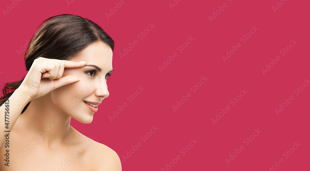 Fototapeta premium Skin care beauty concept. Portrait of smiling woman pointing brow or eye, touching skin, applying face cream, against red color background. Brunette model at studio shot.