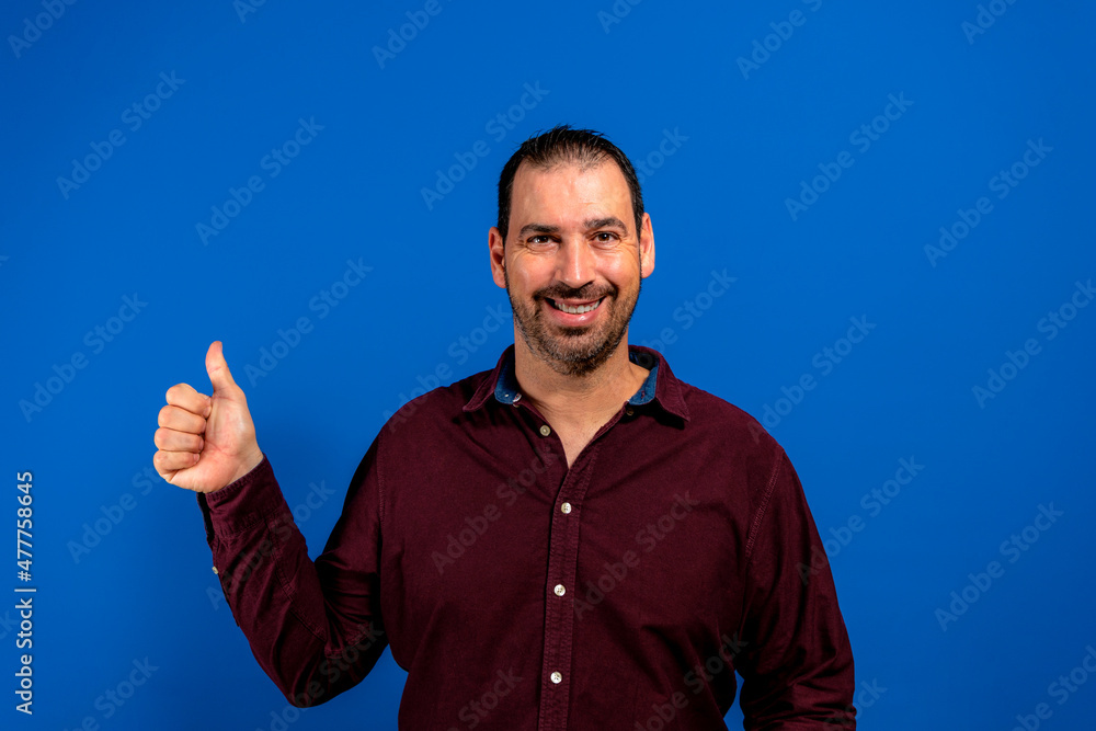 Portrait of cheerful man in basic clothing smiling and showing thumbs up at camera isolated over blue background Portrait of cheerful man in basic clothing smiling and showing thumbs up at camera isolated over blue background