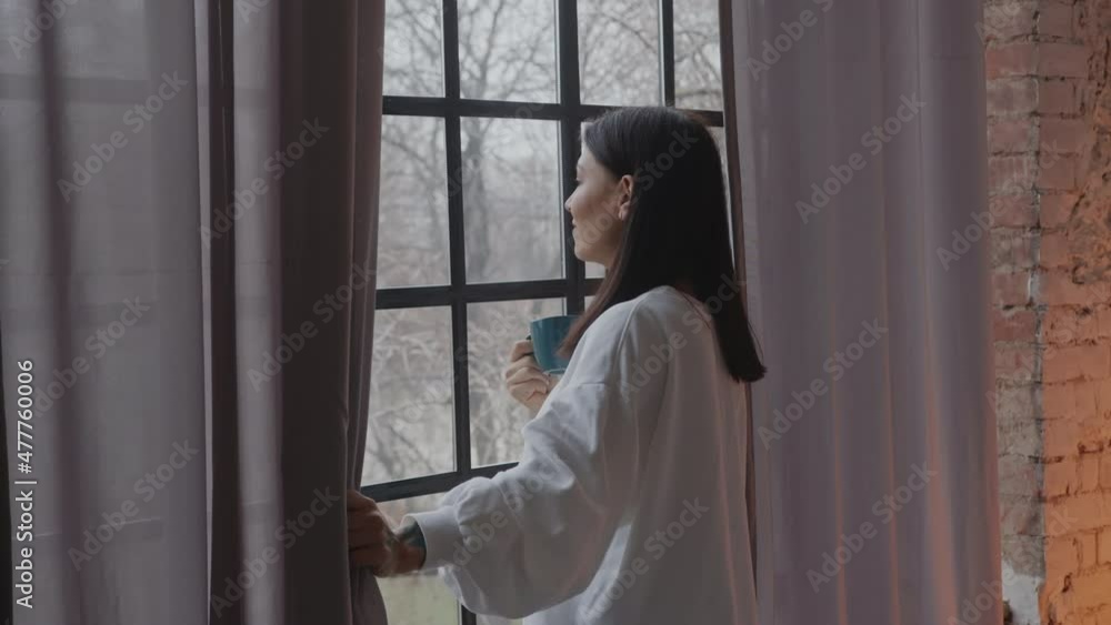 Young caucasian woman drinking morning coffee while looking through the window in winter. High quality 4k footage Young caucasian woman drinking morning coffee while looking through the window in winter. High quality 4k footage