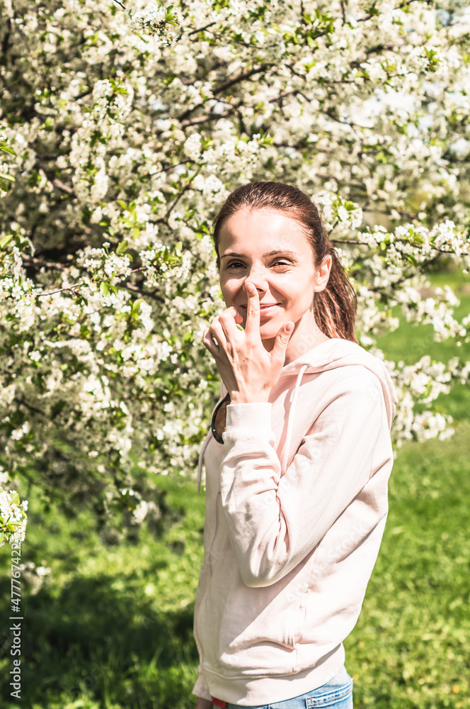 Beautiful young woman posing near spring flowering trees. Girl touches her nose by the finger. Allergy free concept. Model in casual wear posing outdoor in park Beautiful young woman posing near spring flowering trees. Girl touches her nose by the finger. Allergy free concept. Model in casual wear posing outdoor in park