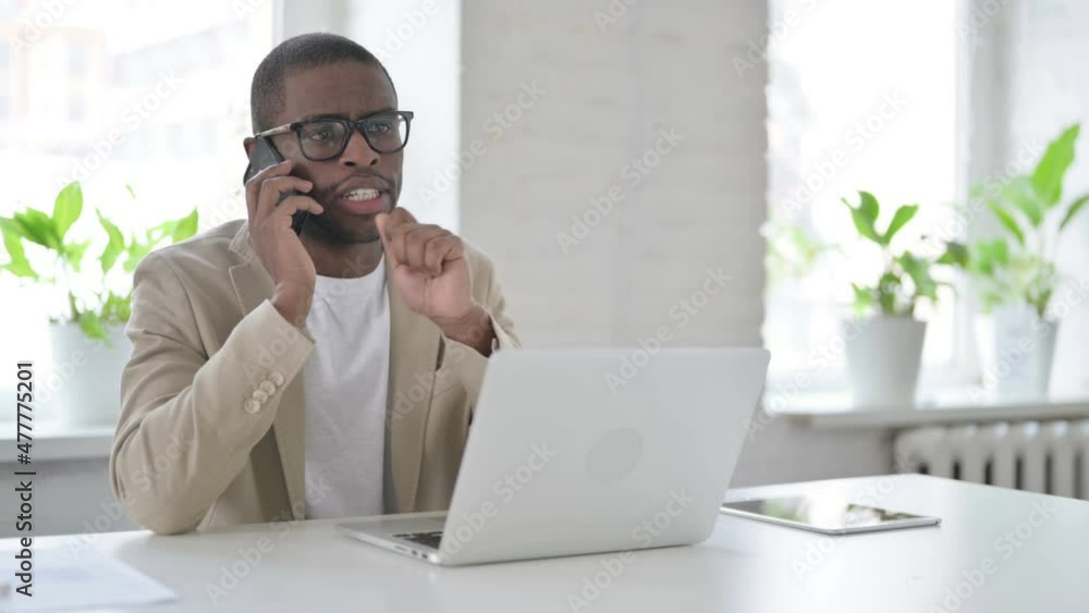 African Man Talking on Smartphone while using Laptop in Office African Man Talking on Smartphone while using Laptop in Office
