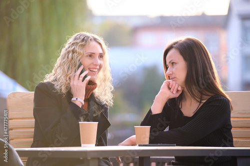 Unhappy young woman waiting angrily while her friend is talking happily on sellphone with someone else and ignoring her. Friendship problems concept