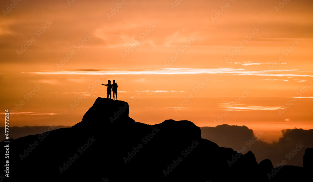 Dramatic golden sunset of two men admiring the view on vacation, standing on a large rock on a beach in Jacobsbaai, South Africa.