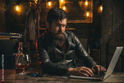 Stylish rich man holding a glass of old whisky. Bearded man enjoying whiskey or cognac and doing his business at laptop.