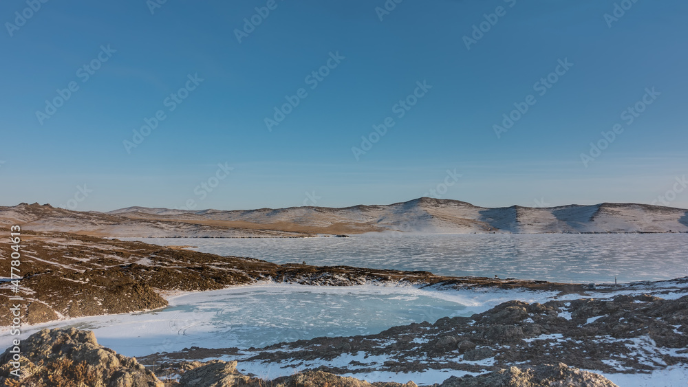 A small frozen heart-shaped lake is separated from a large one by a strip of land. There are snow patterns and tire tracks on the ice. Hills against a blue sky. Snow on the banks. Baikal
