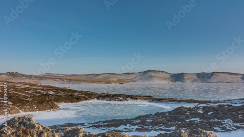 A small frozen heart-shaped lake is separated from a large one by a strip of land. There are snow patterns and tire tracks on the ice. Hills against a blue sky. Snow on the banks. Baikal