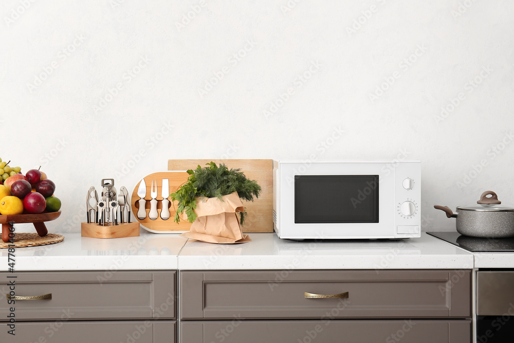 Modern microwave oven, kitchenware and food on counter near light wall Modern microwave oven, kitchenware and food on counter near light wall