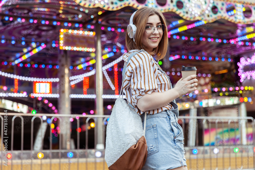 Cute stylish woman in glasses and denim overalls, listening to music on headphones, holding coffee cup in an amusement park