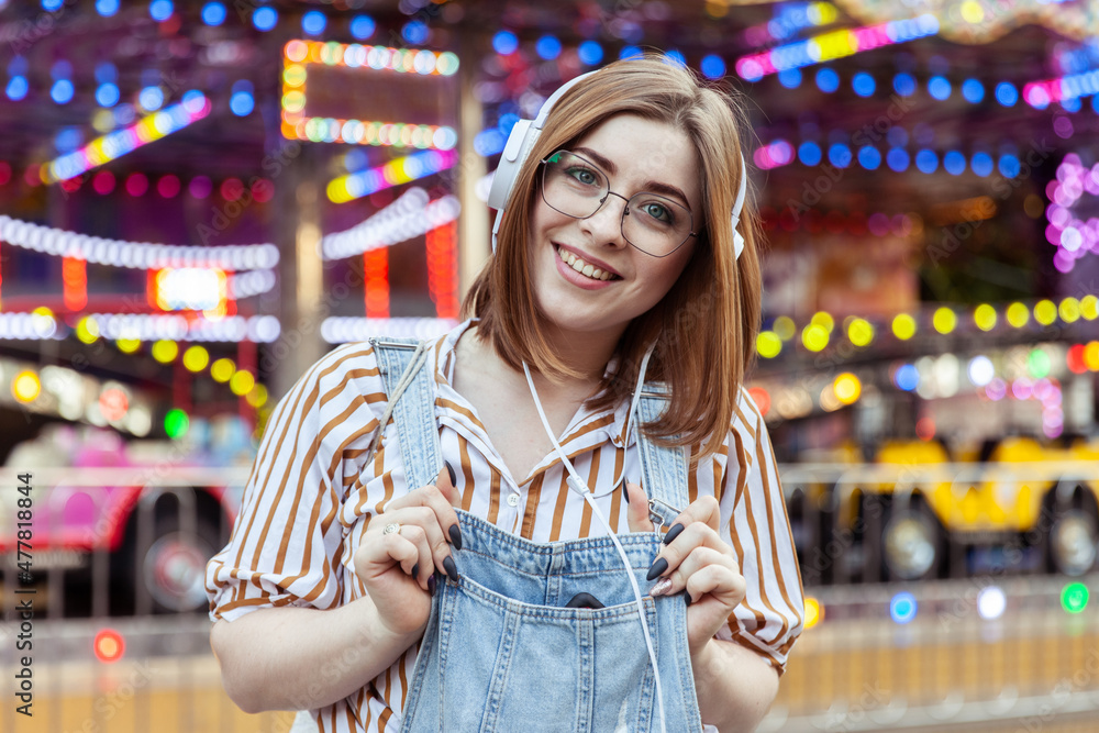 Cute smiling woman in denim overalls and glasses listens to music on headphones in an amusement park