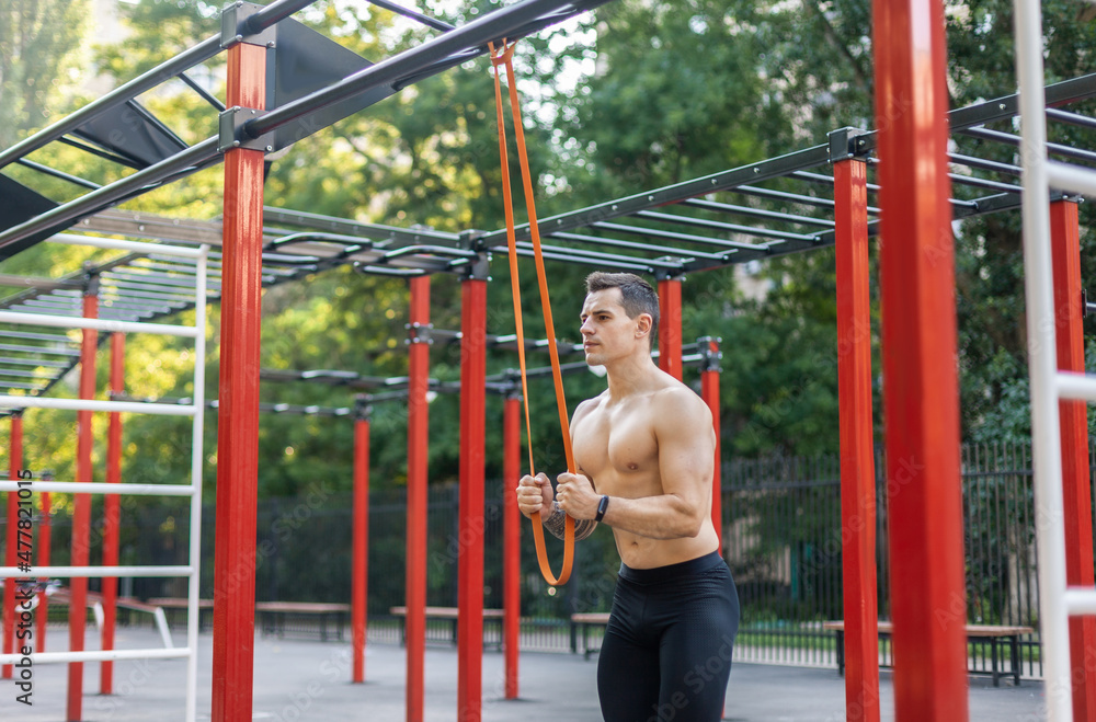 Athletic male athlete with a naked torso trains with a fitness elastic band on the workout site. Outdoor workout