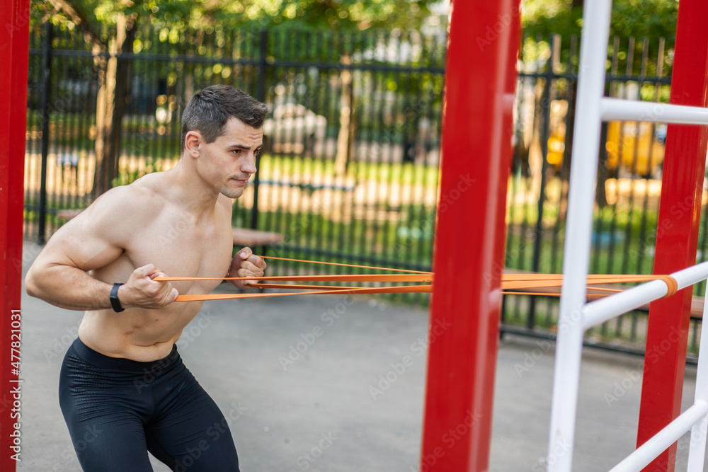 Obraz premium Athletic male athlete with a naked torso trains with a fitness elastic band on the workout site. Outdoor workout