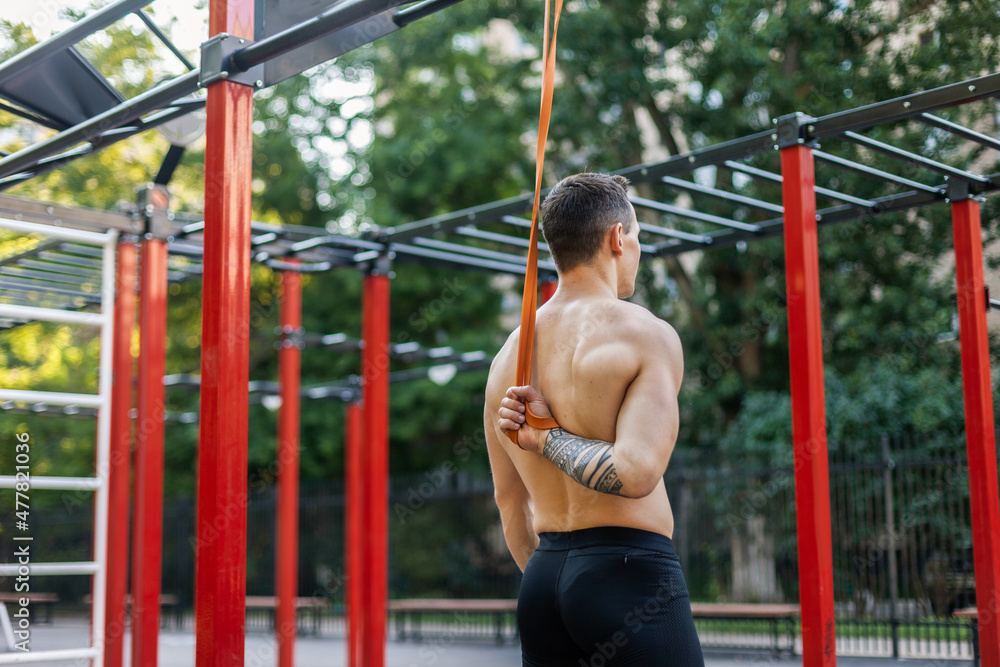 Athletic male athlete with a naked torso trains with a fitness elastic band on the workout site. Outdoor workout