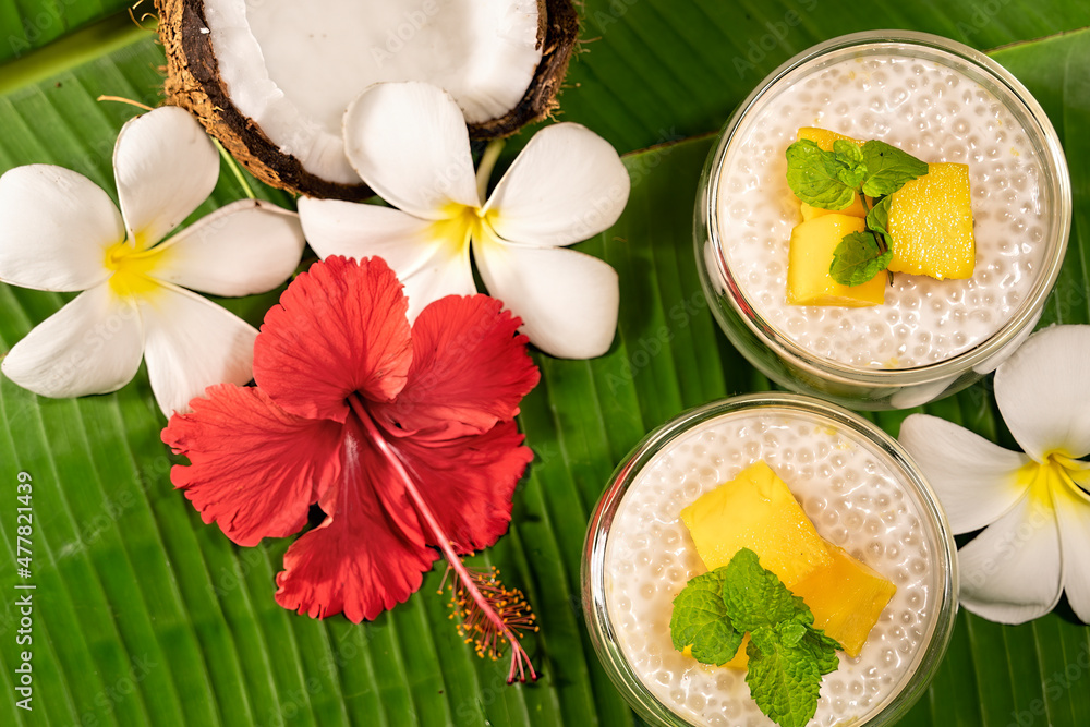 Tropical tapioca and coconut cream with mango dessert on green banana leaf background  frangipani and hibiscus flowers flat lay.