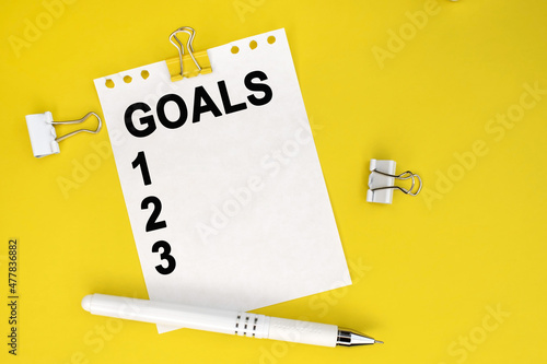 The inscription of the goal is written on a white piece of paper from a notepad that lies on a yellow background near a pen and paper clips.