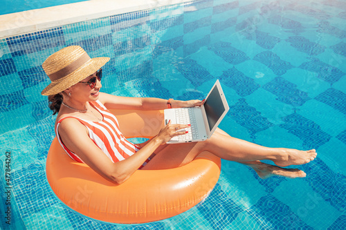 Woman in a swimsuit floating in inflatable circle in swimming pool and surfing the internet websites at her laptop pc. Leisure weekend and remote freelance work concept