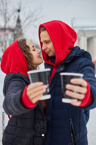 young family guy and girl spend the day in the park on a snowy day. the guy hugs the girl while standing on the street, they drink coffee together.