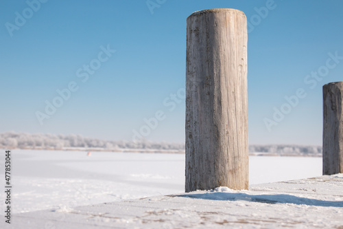 Detailaufnahme von hölzernen Stegpfählen am Wallendorfer See im Winter mit Schnee und Eis. Das Motiv zeigt Ruhe, Kälte und reduzierte Landschaftsstimmung in natürlicher Umgebung.
