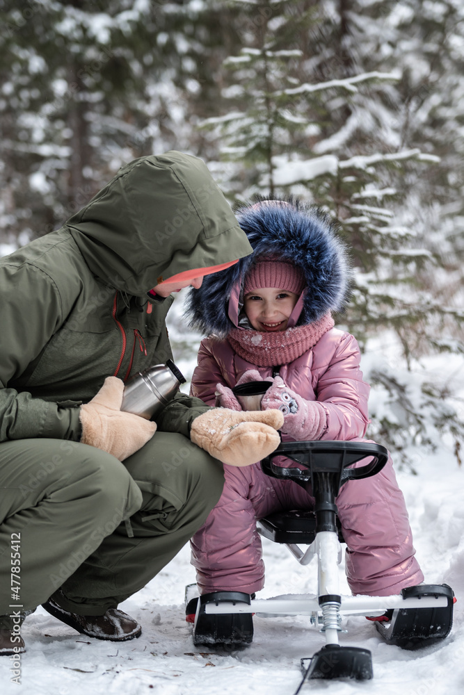 Family outdoor activities. Mom and daughter are resting and drinking tea after an active walk in the winter forest.