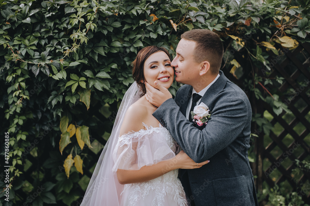 Wedding portrait of a stylish groom and a beautiful, smiling brunette girl with a bouquet in her hands. Lovely newlyweds hug on a background of green ivy foliage in nature. Wedding portrait of a stylish groom and a beautiful, smiling brunette girl with a bouquet in her hands. Lovely newlyweds hug on a background of green ivy foliage in nature.