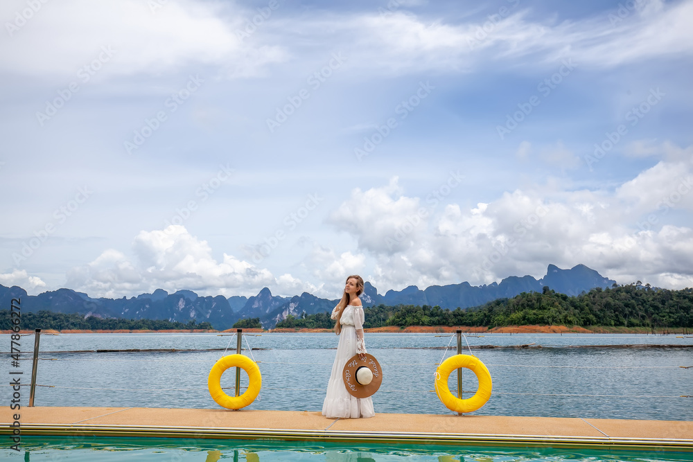 Happy Travel Girl with Hat Fun on Wooden Pier with Lake, Rainforest Jungle and Mountains on Background. Female Tourist Enjoy Vacation Cheow Lan Lake in Khao Sok National Park in Thailand
